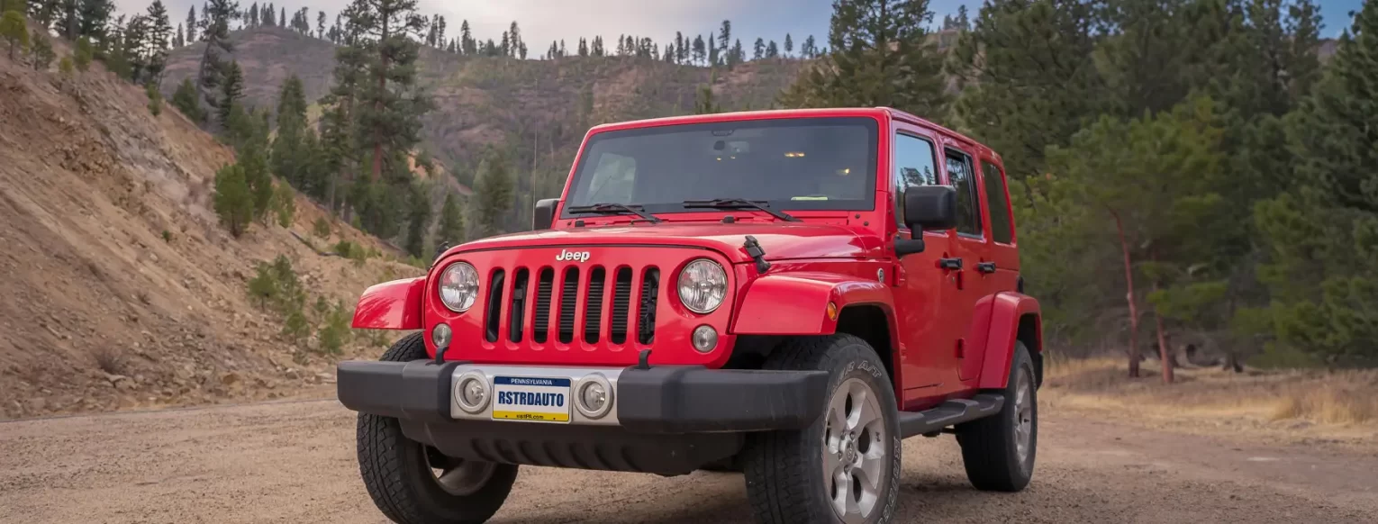 A red Jeep on a dirt road with trees and mountains behind it.